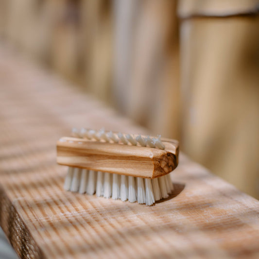 A small nail brush with a light wood handle and white bristles resting on a wooden surface in warm natural light.
