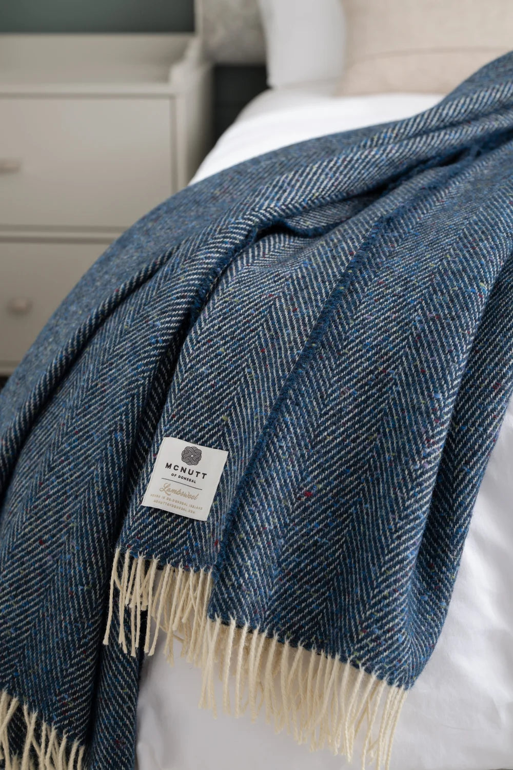 McNutt Arranmore blue Donegal lambswool tweed throw draped across a bed, showing herringbone weave, fringe and label