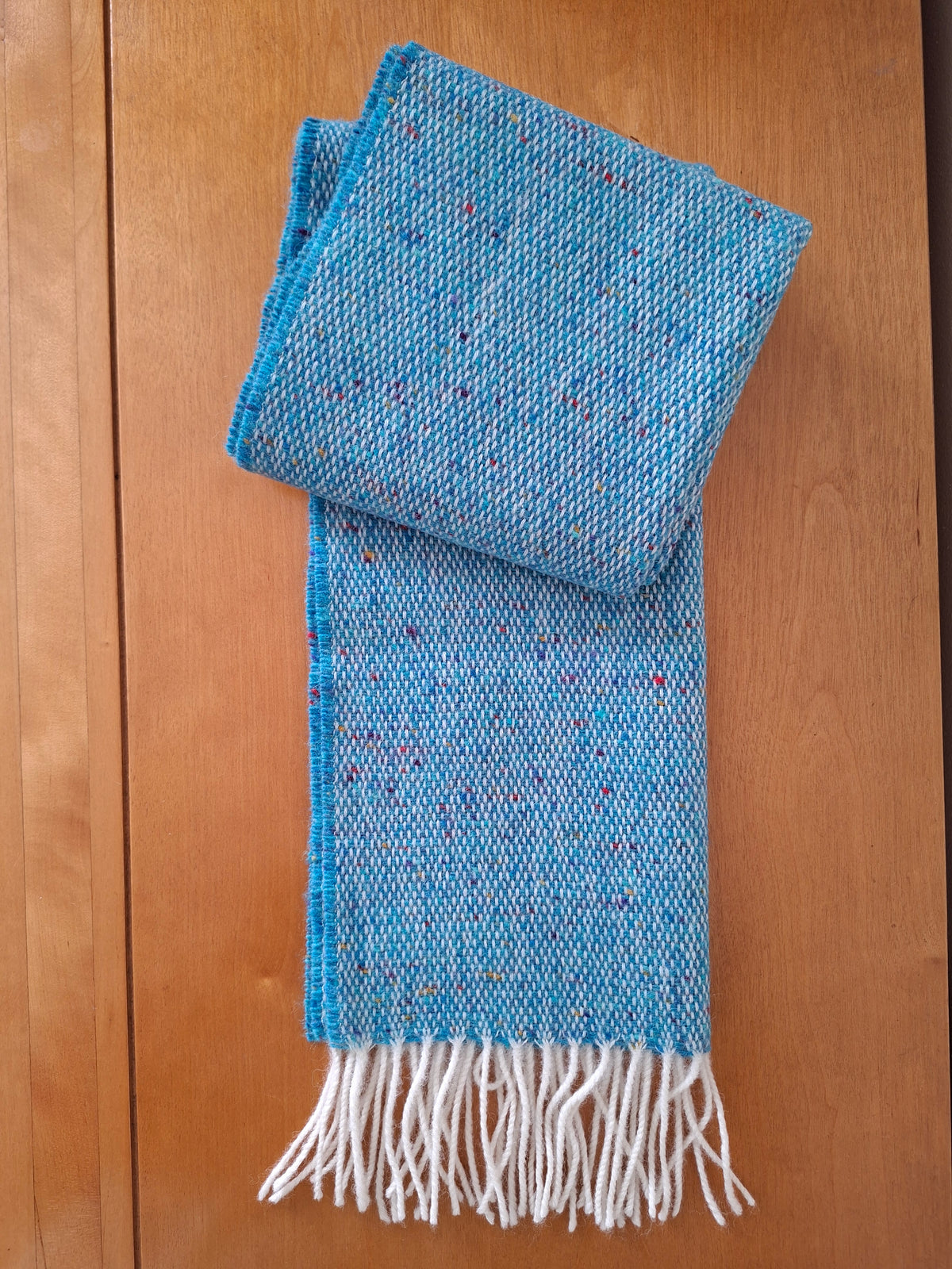McNutt Donegal lambswool tweed scarf in peacock with off white fringe, folded on wooden background.