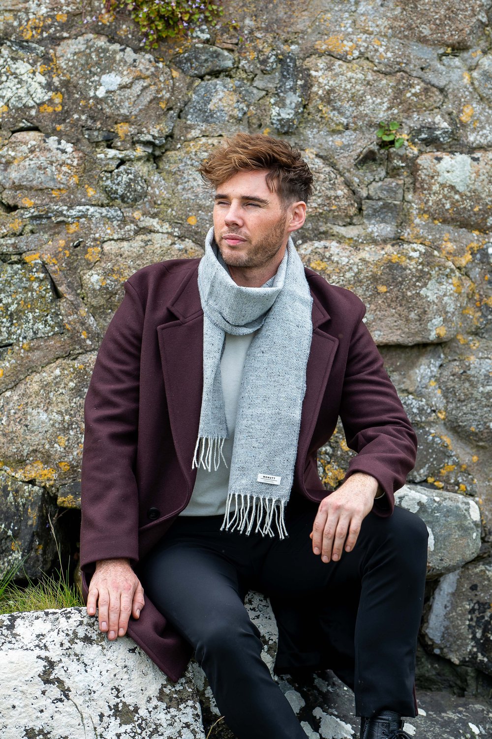 Gentleman wearing McNutt Donegal lambswool tweed scarf in Donkey Grey sitting on rocks on natural Donegal landscape. 