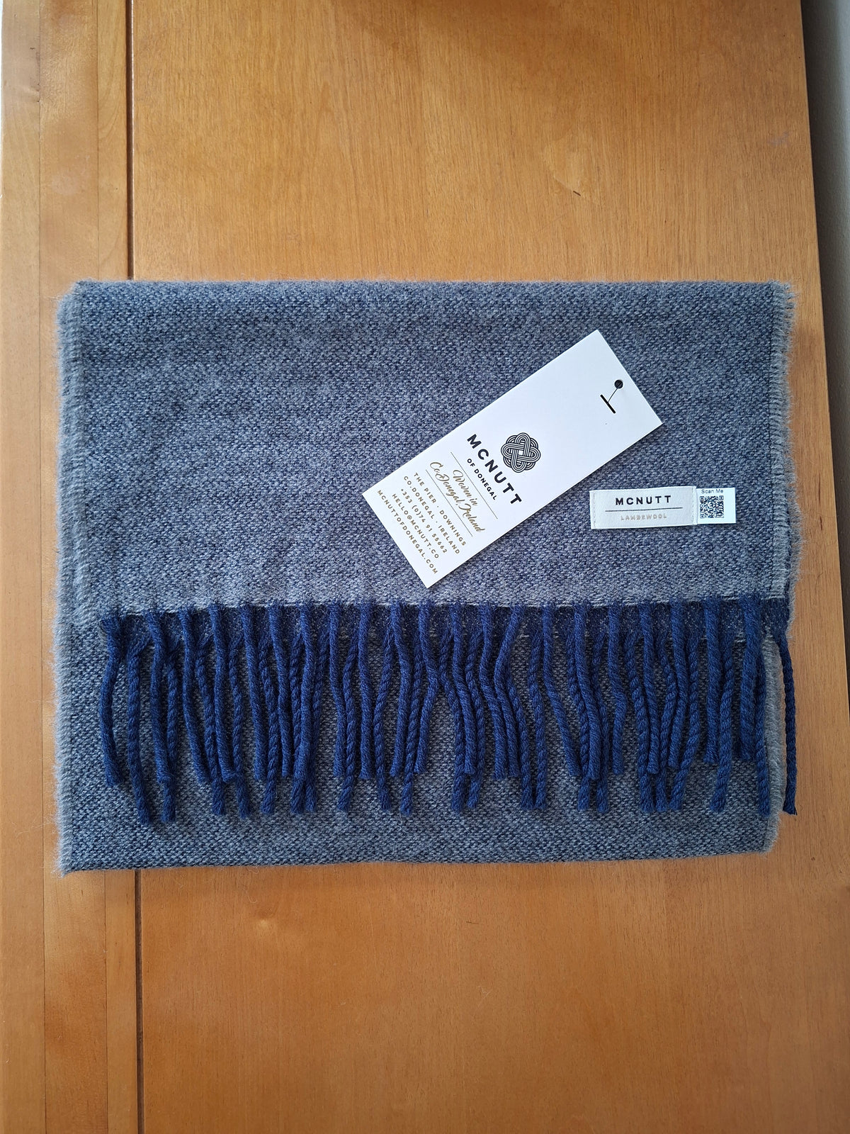 A folded lambswool scarf with dark blue fringe, laid flat on a wooden surface. A white hangtag with the brand name McNutt is attached to the scarf.