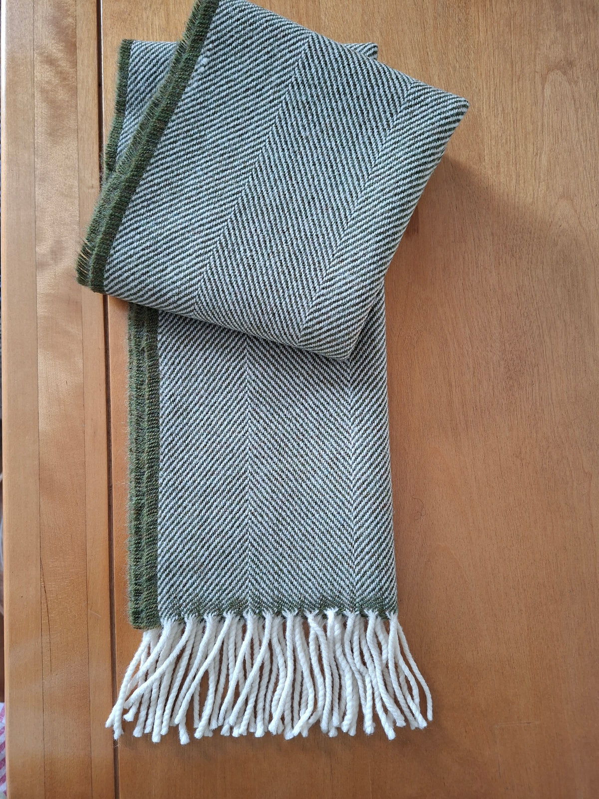 A folded lambswool herringbone-patterned scarf in sage green and white, with a solid dark green border and white fringe tassels, laid flat on a wooden surface.