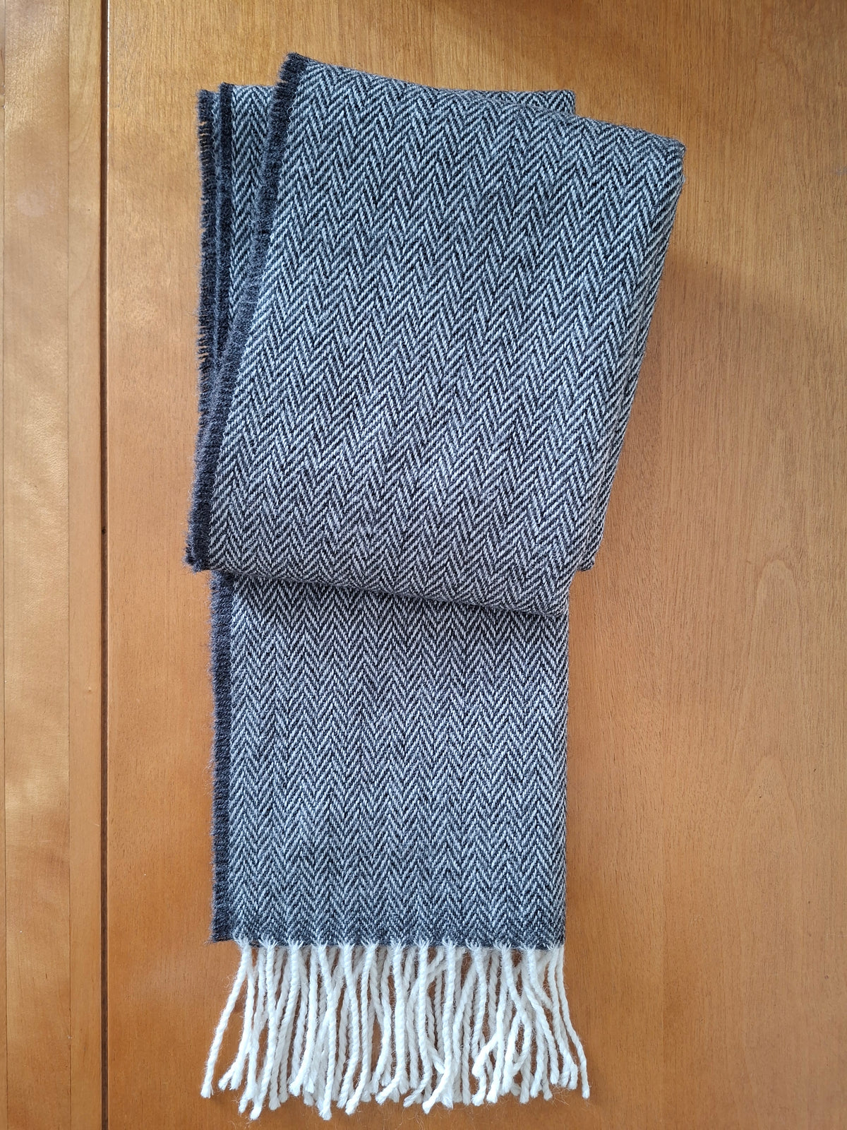 A folded herringbone pattern scarf in slate blue and white with white fringe tassels, laid flat on a wooden surface.
