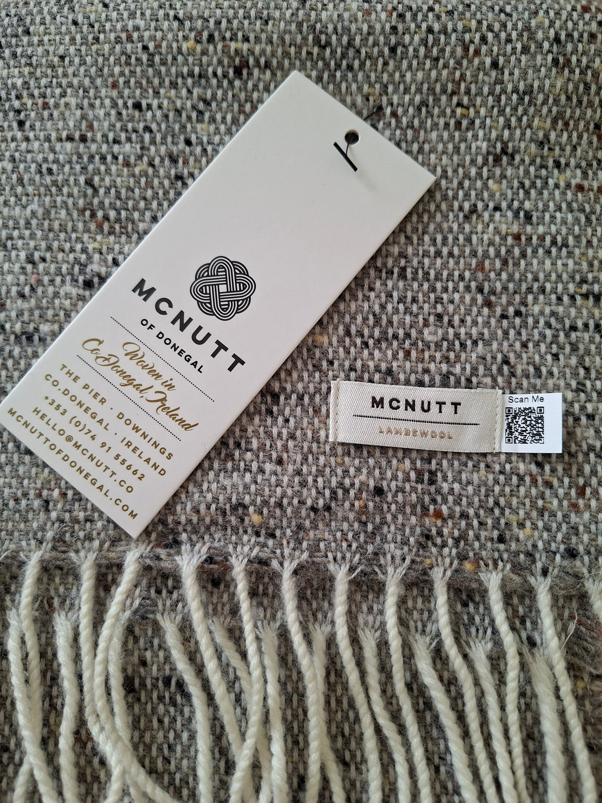 A close-up of lambswool donkey grey scarf with cream fringe, showing a white brand tag that reads McNutt of Donegal and a small woven label.