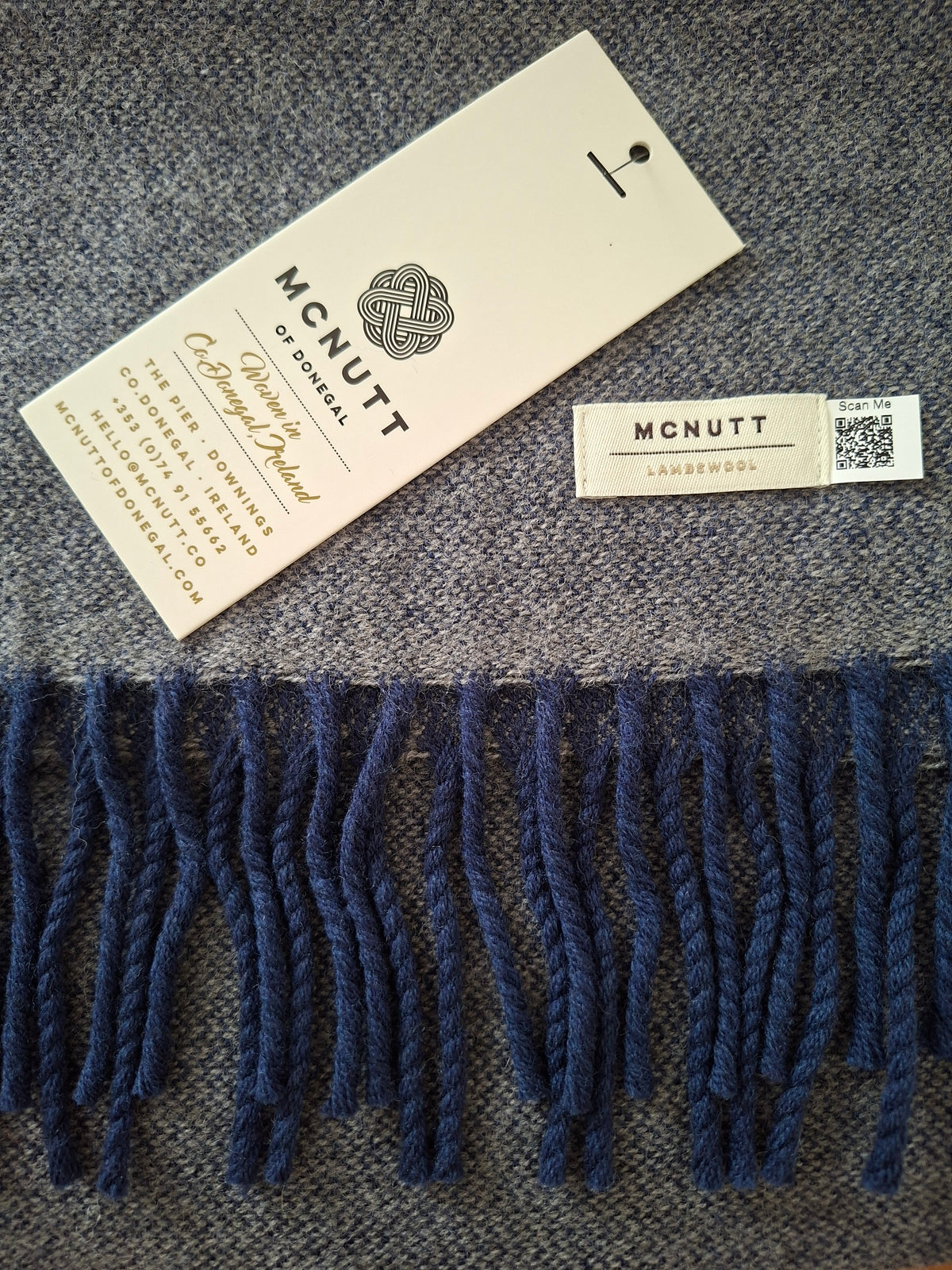 A close-up of a woven scarf with dark navy blue fringe, laid on a textured gray fabric background. The scarf includes a cream paper hangtag and a sewn-in label both printed with the brand name MCNUTT.