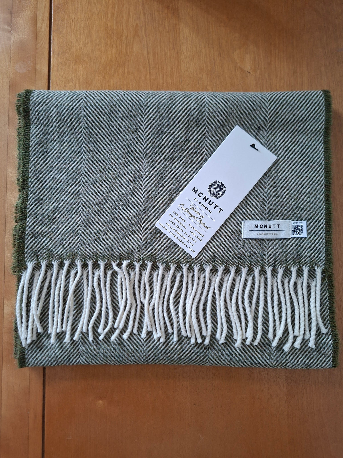 A folded lambswool scarf in a green herringbone pattern with grey white fringe, resting on a wooden surface with its tags attached.