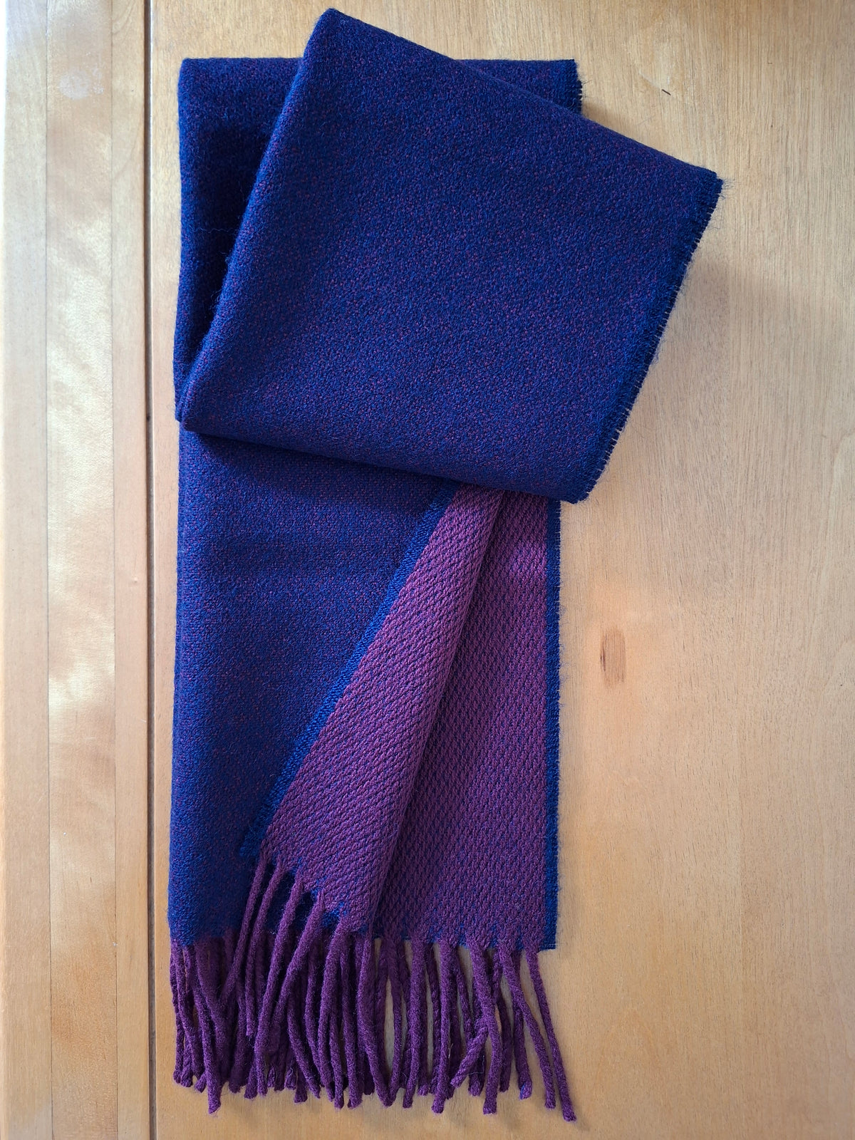 A folded woven scarf in two colors: a dominant deep navy blue and a secondary burgundy section with matching fringe, displayed on a light wood background.