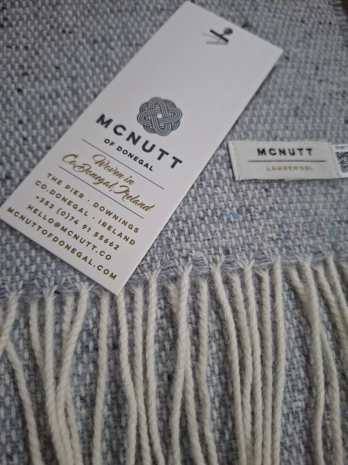 A close-up of lambswool light grey scarf with cream fringe, showing a white brand tag that reads McNutt of Donegal and a small woven label.