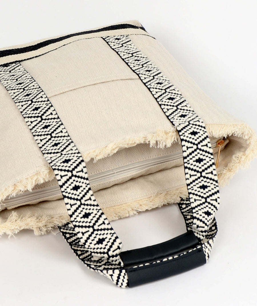 Top of bag view of off-white canvas tote bag by Pia Rossini with zipped closure, fringed accents and black geometric pattern on handles against a white background
