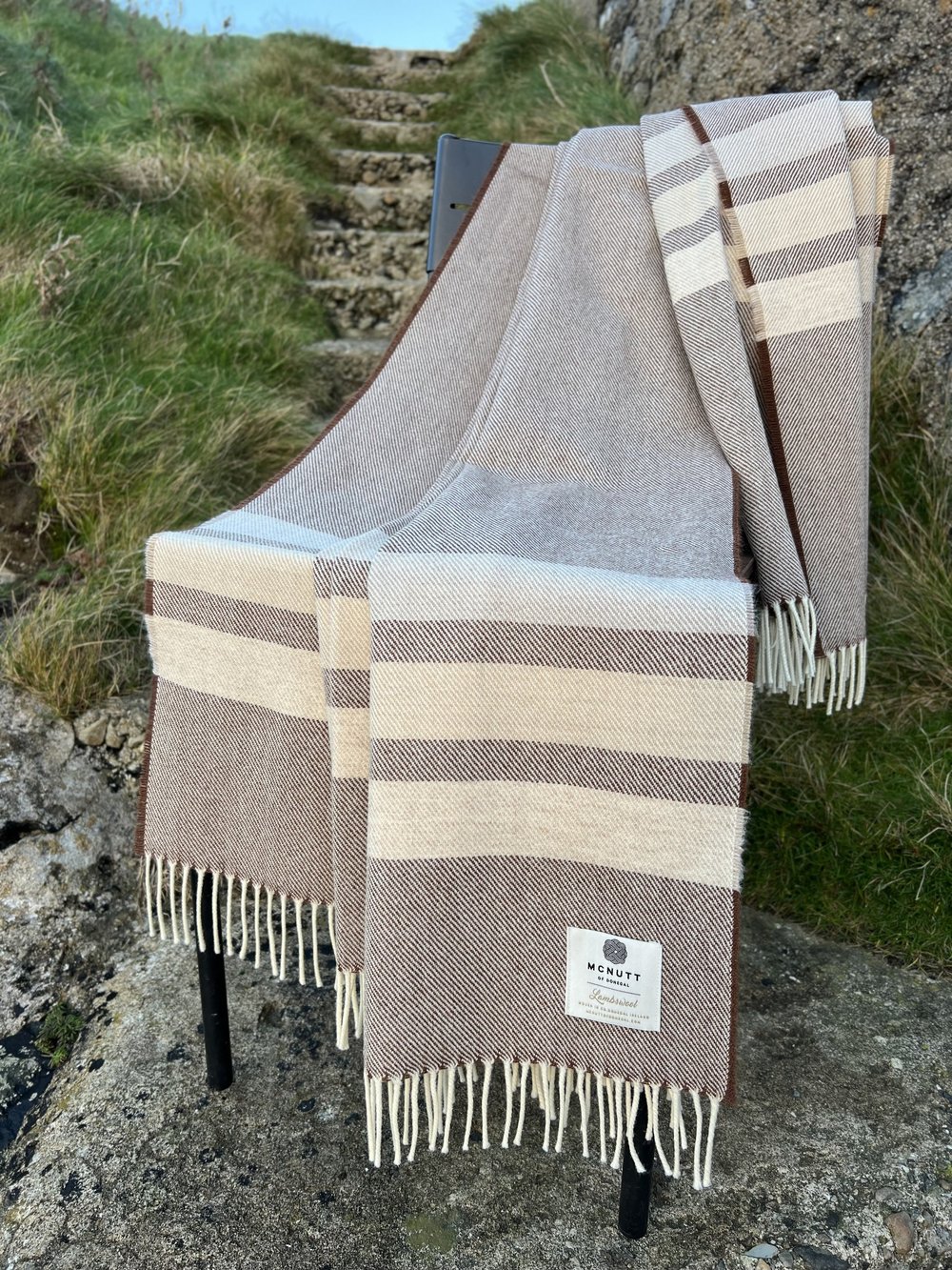 McNutt of Donegal Coffee Brew Merino lambswool throw displayed outdoors, showing brown and cream bands and fringed edges