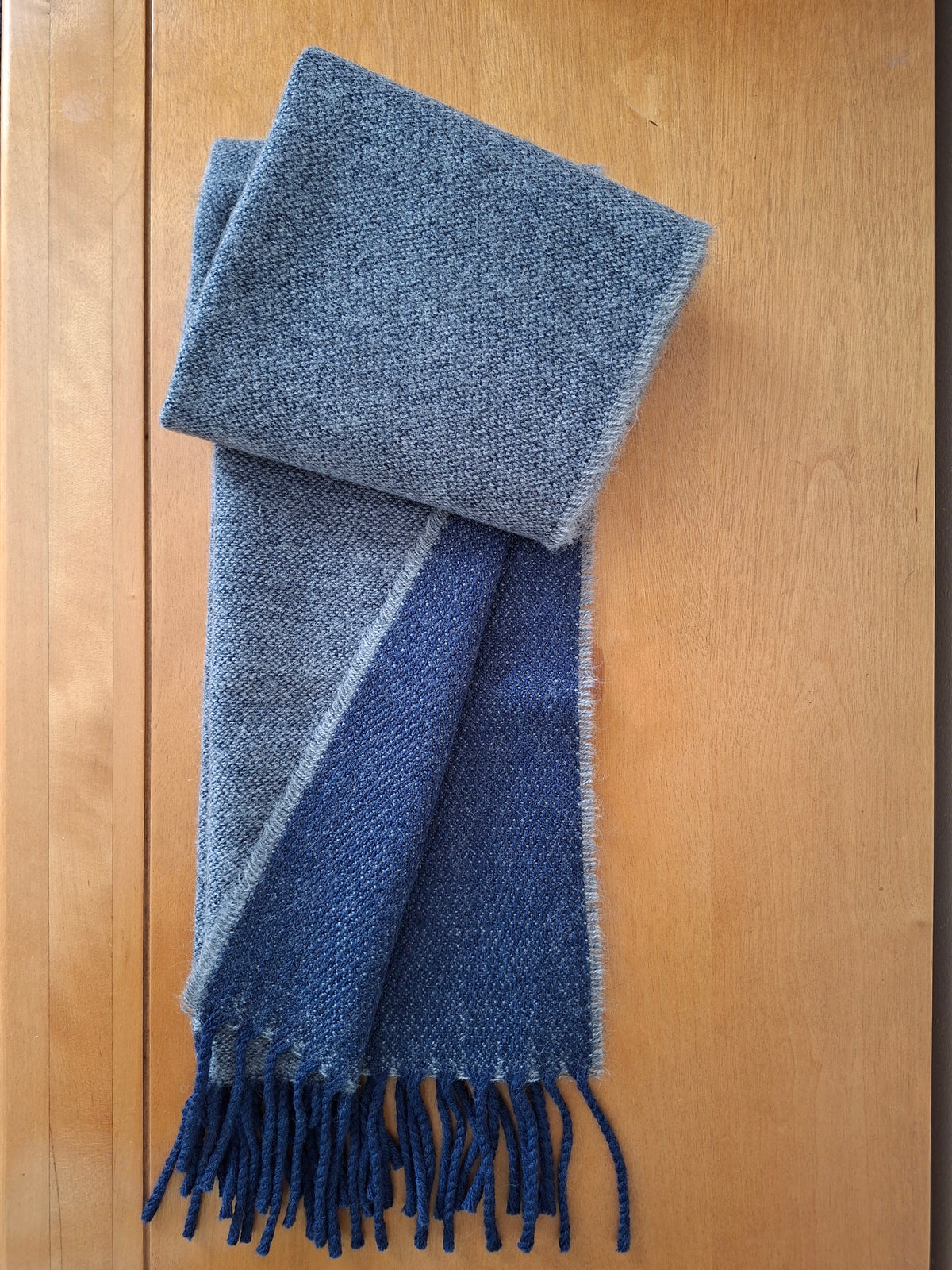 A folded lambswool scarf with a heathered slate blue outer section and a solid dark blue inner section with fringe, displayed on a light wood background.