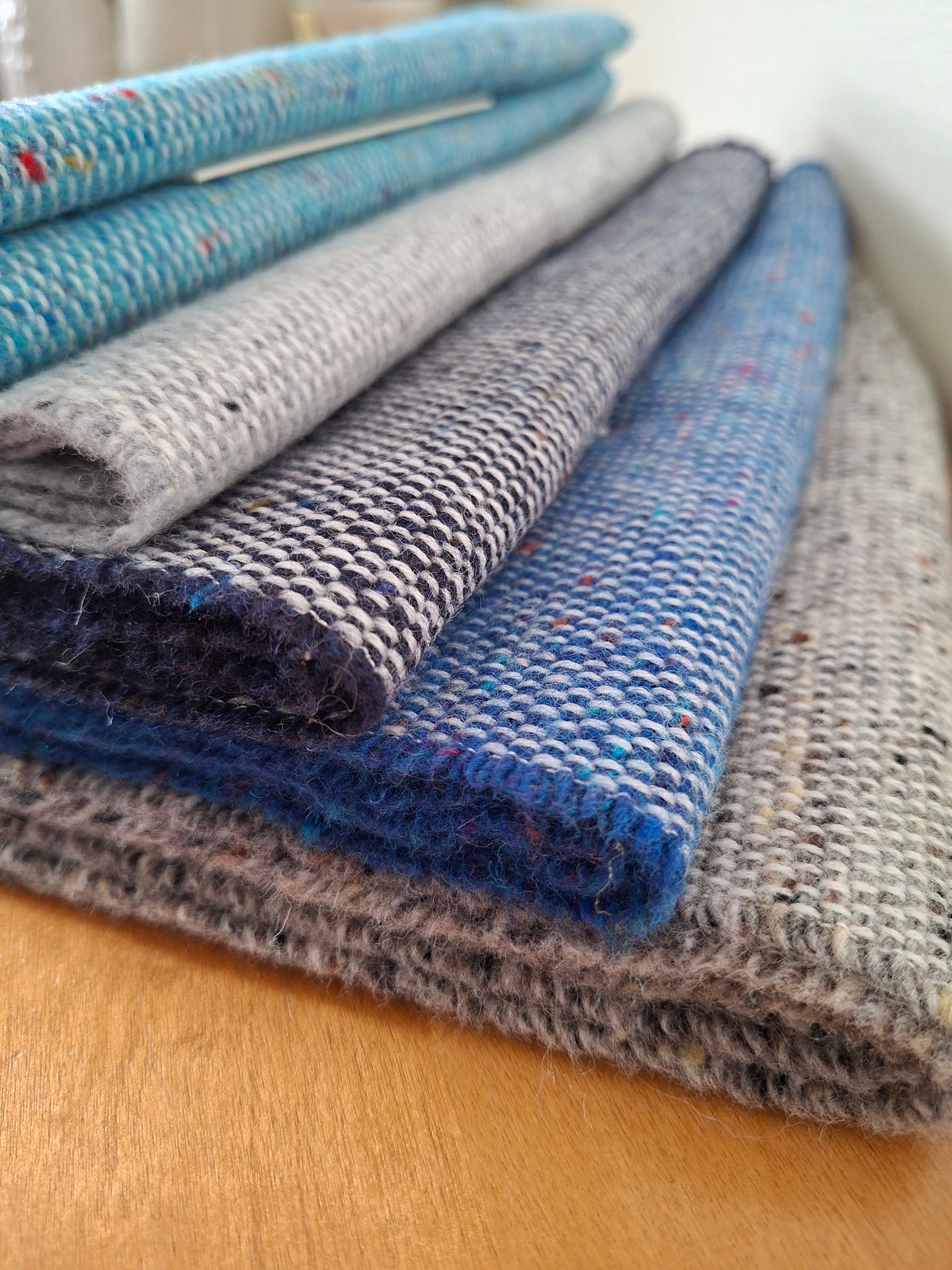 A stack of folded McNutt's lambswool scarves in blue and grey tones with small multicoloured flecks, photographed on a wooden surface.