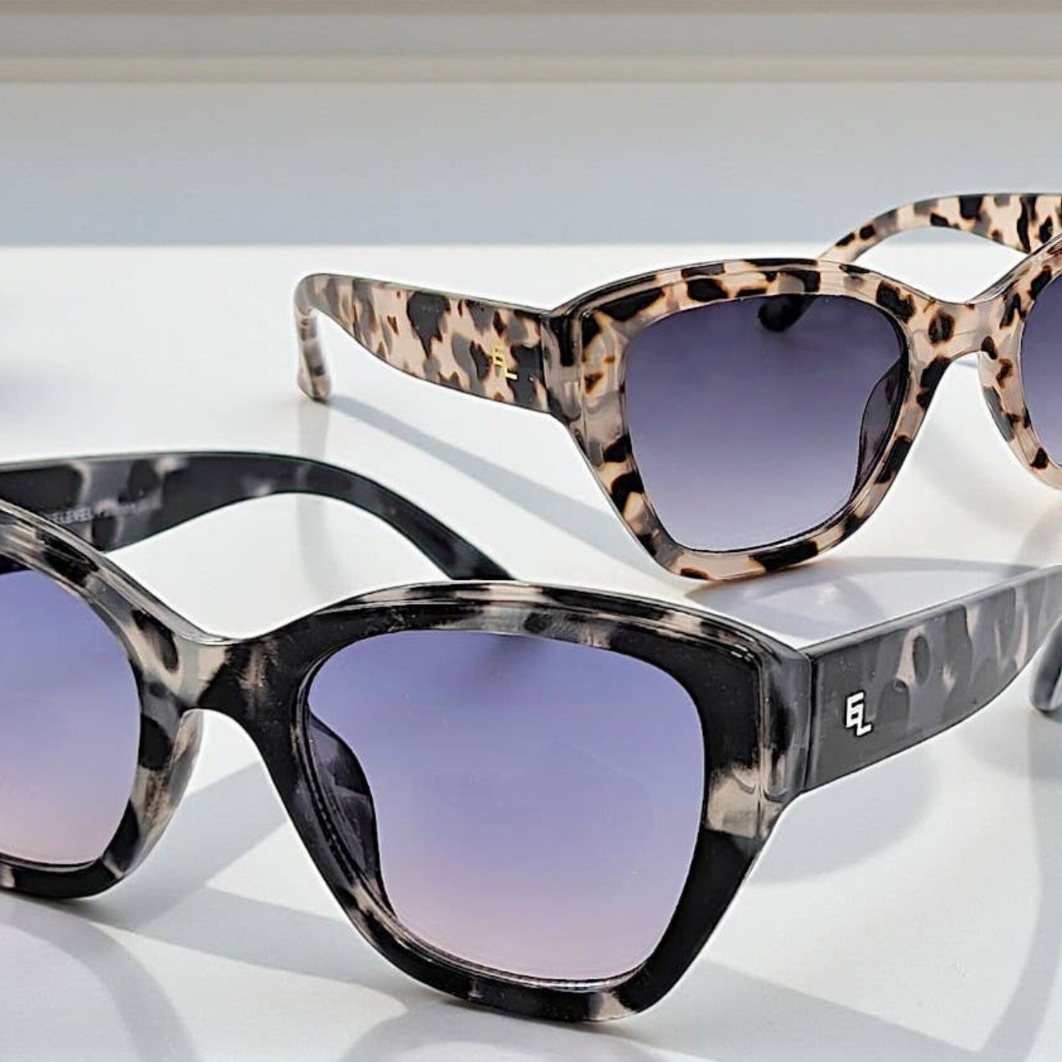 Two pairs of EyeLevel Grace ladies fashion sunglasses displayed on a white surface, featuring bold square frames with gradient lenses—one in black marble and one in tortoiseshell, styled in natural light