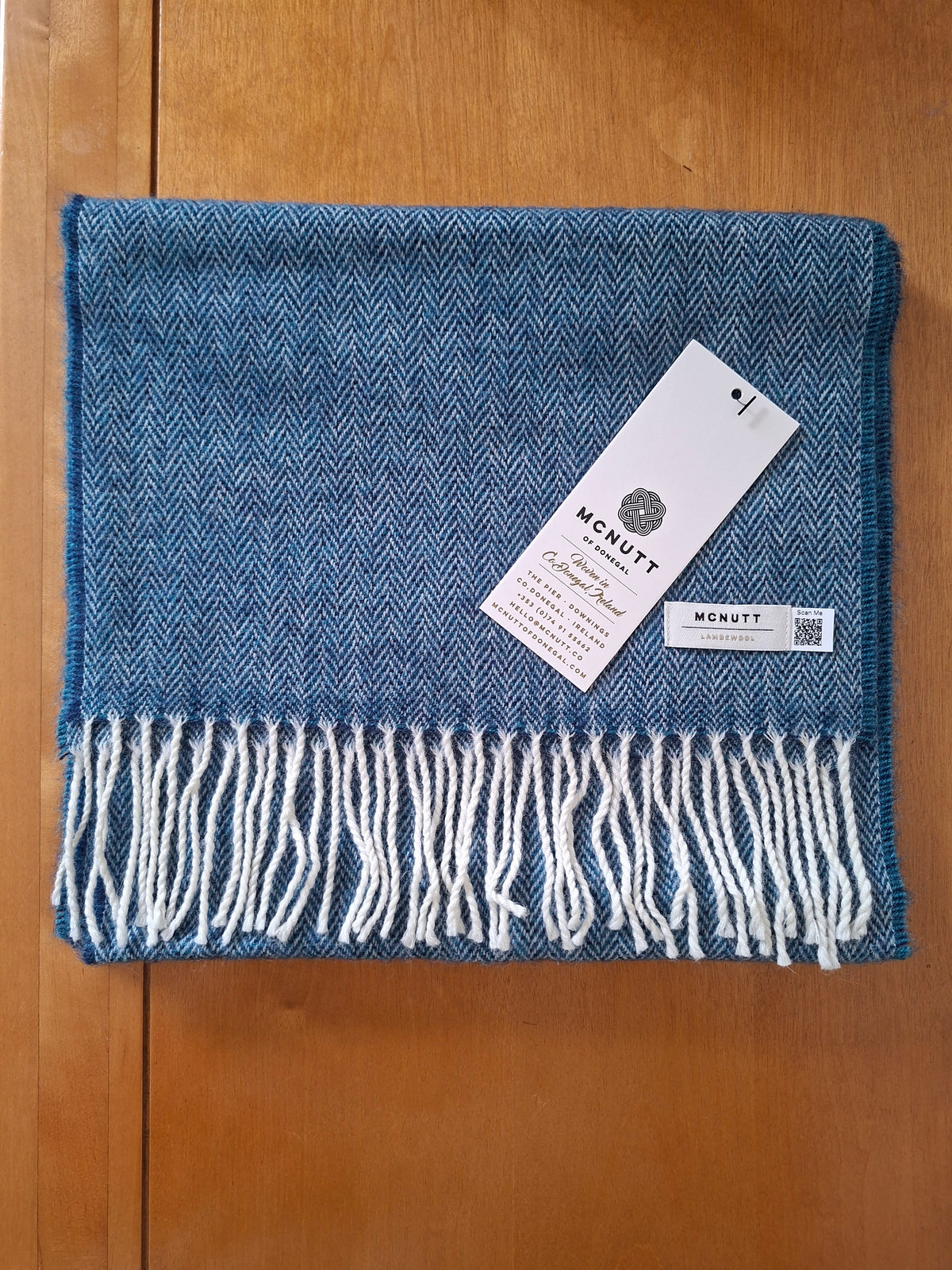 A folded blue lambswool herringbone woven scarf with grey white fringe, resting on a wooden surface. A white brand tag for 'McNutt' is attached to the scarf.