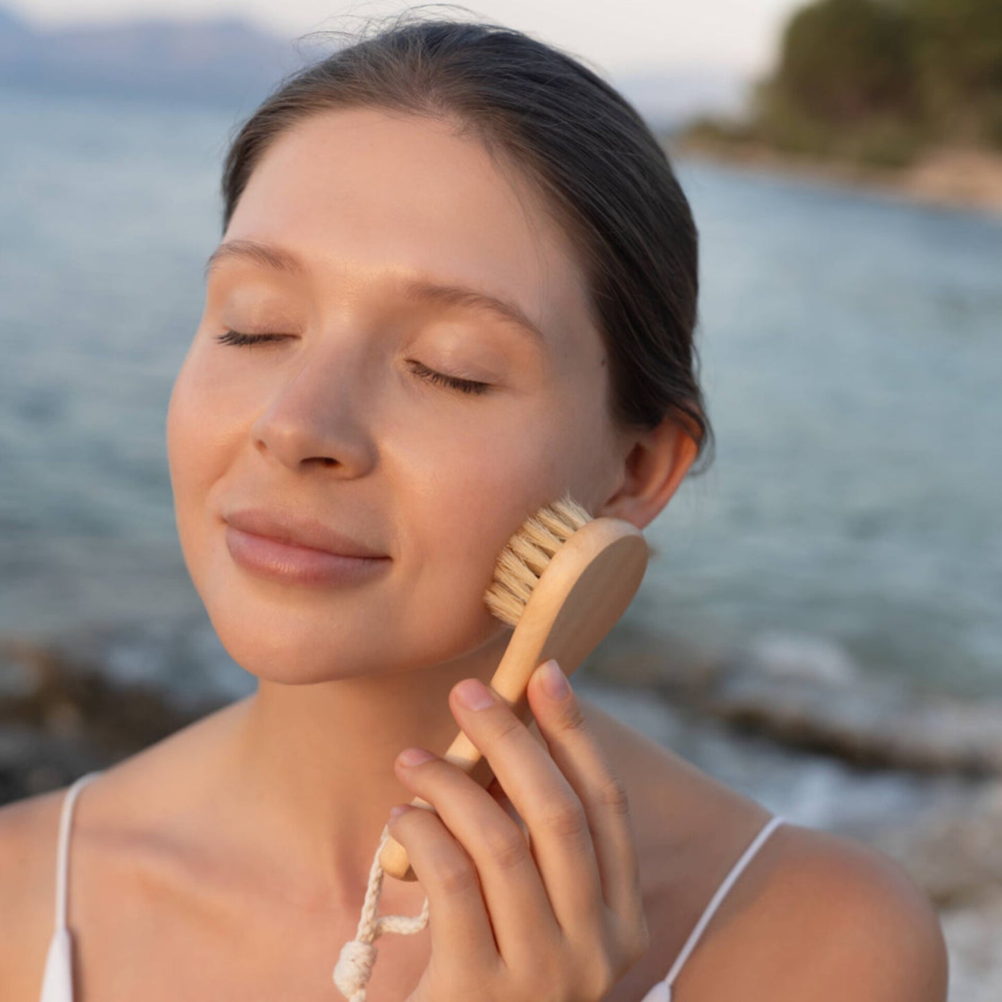 Woman using a Croll and Denecke facial cleansing wooden handled brush on her face with a blurred natural background