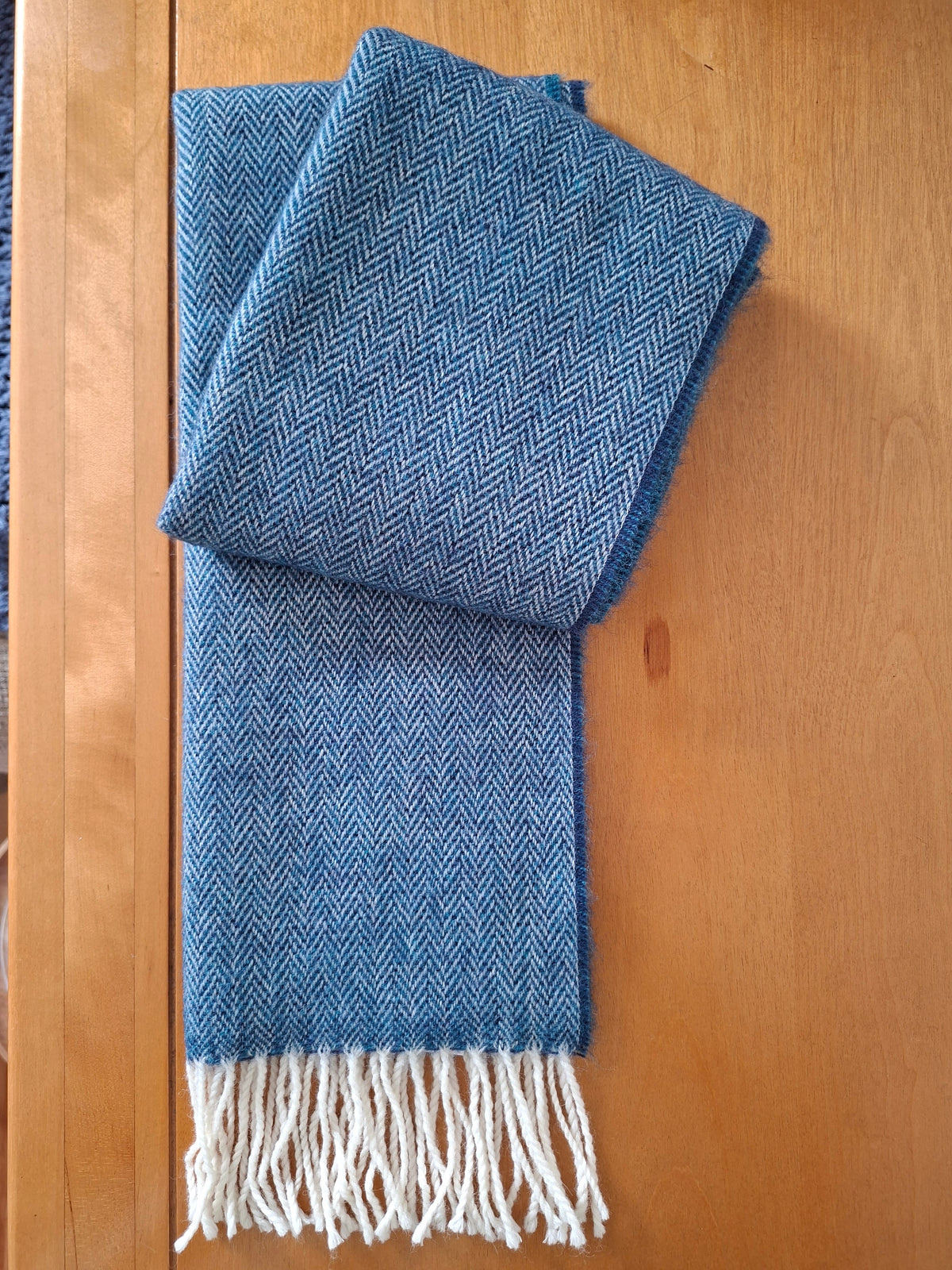 A folded blue herringbone patterned scarf with grey white fringe tassels, laid flat on a light brown wooden surface.