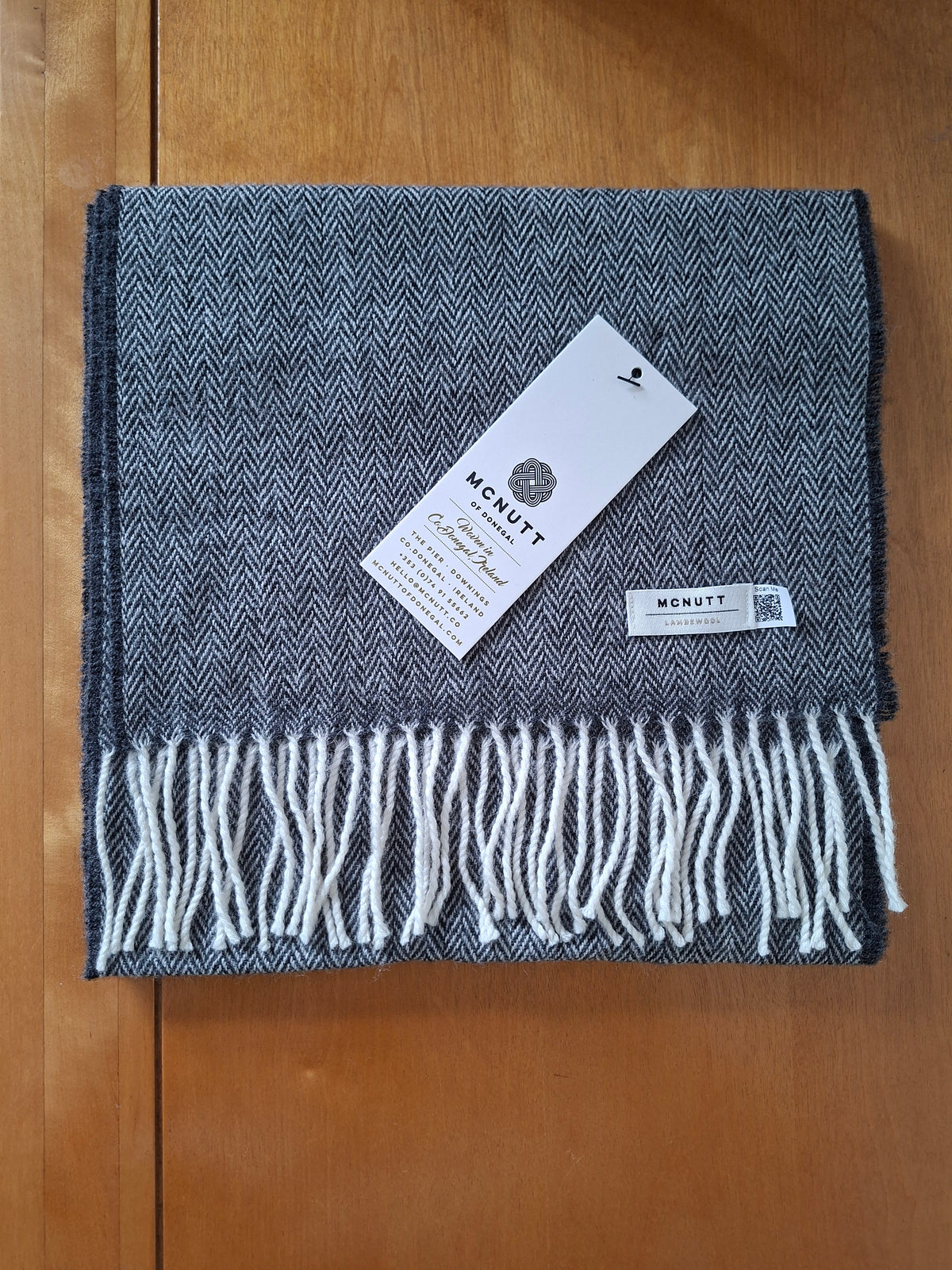 A folded gray and white herringbone pattern scarf with white fringe, featuring a white paper tag with the brand name McNuttplaced on top, set against a wooden background.