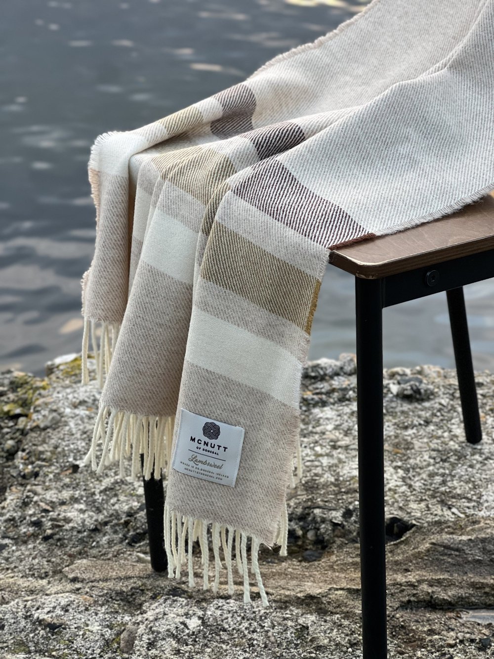 McNutt of Donegal Toffee Roast Merino lambswool throw displayed beside the water, showing beige neutral bands and fringed edges
