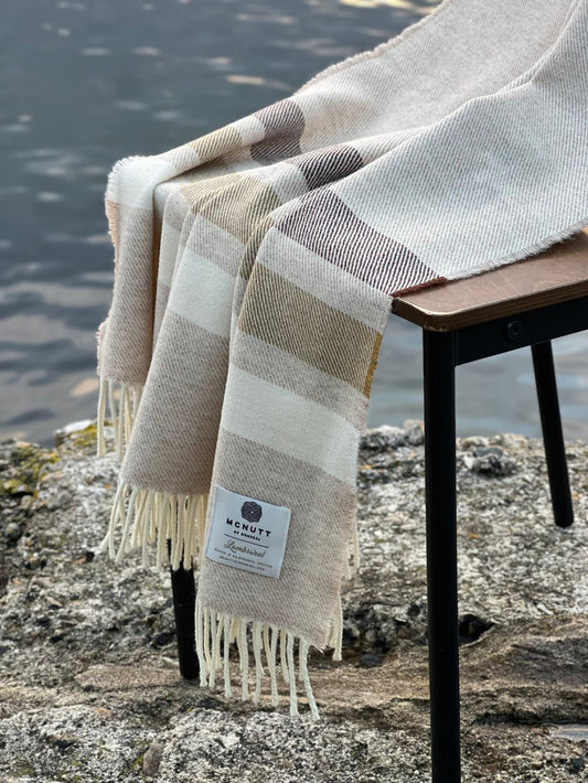 McNutt of Donegal Toffee Roast Merino lambswool throw displayed beside the water, showing beige neutral bands and fringed edges