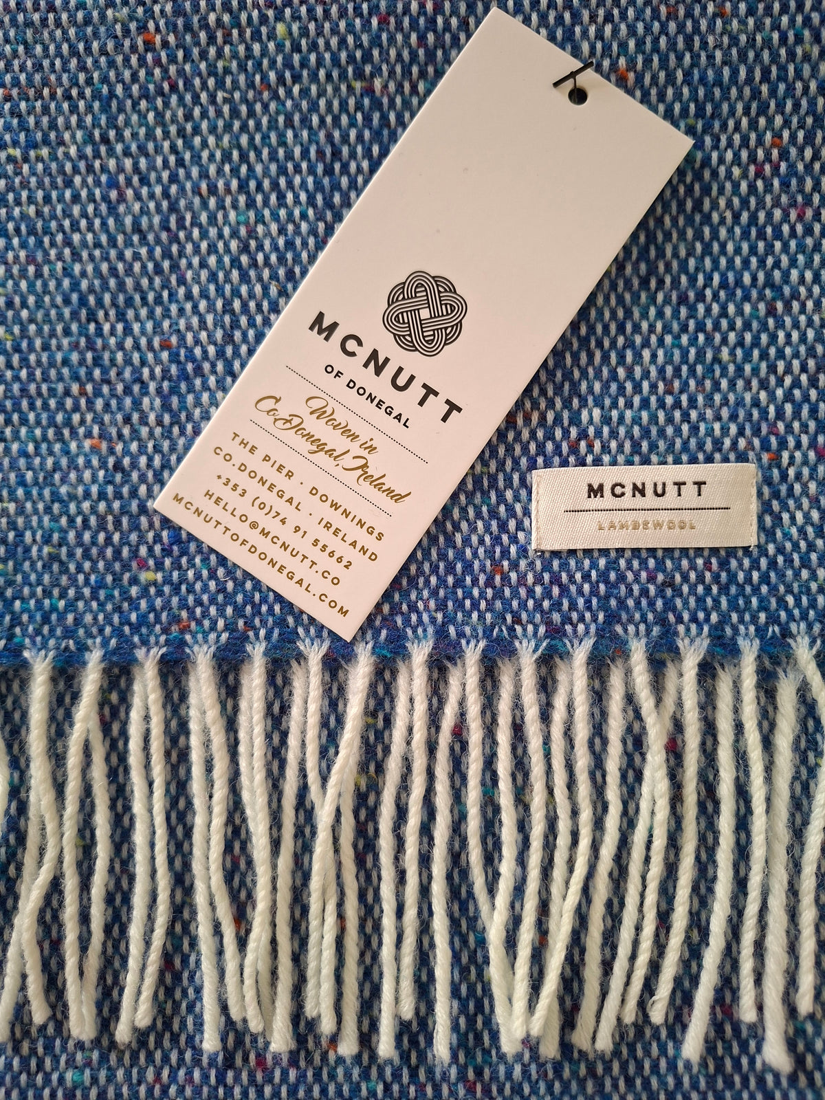 A close-up of lambswool scarf in ocean blue with cream fringe, showing a white brand tag that reads McNutt of Donegal and a small woven label.