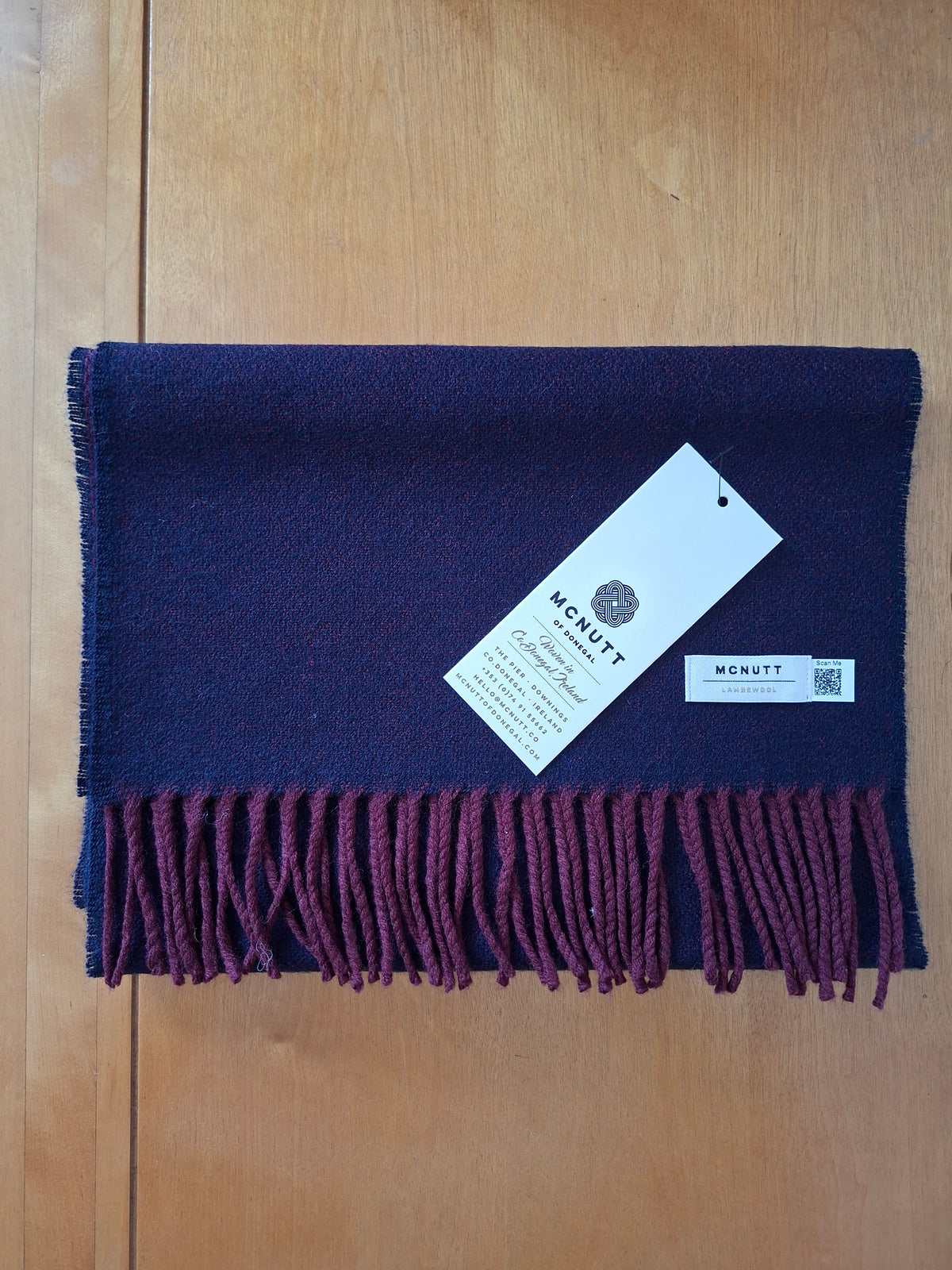 A folded McNutt Lambswool scarf in deep navy blue with a contrasting dark burgundy fringe, displayed on a light wood background with its paper tags attached.