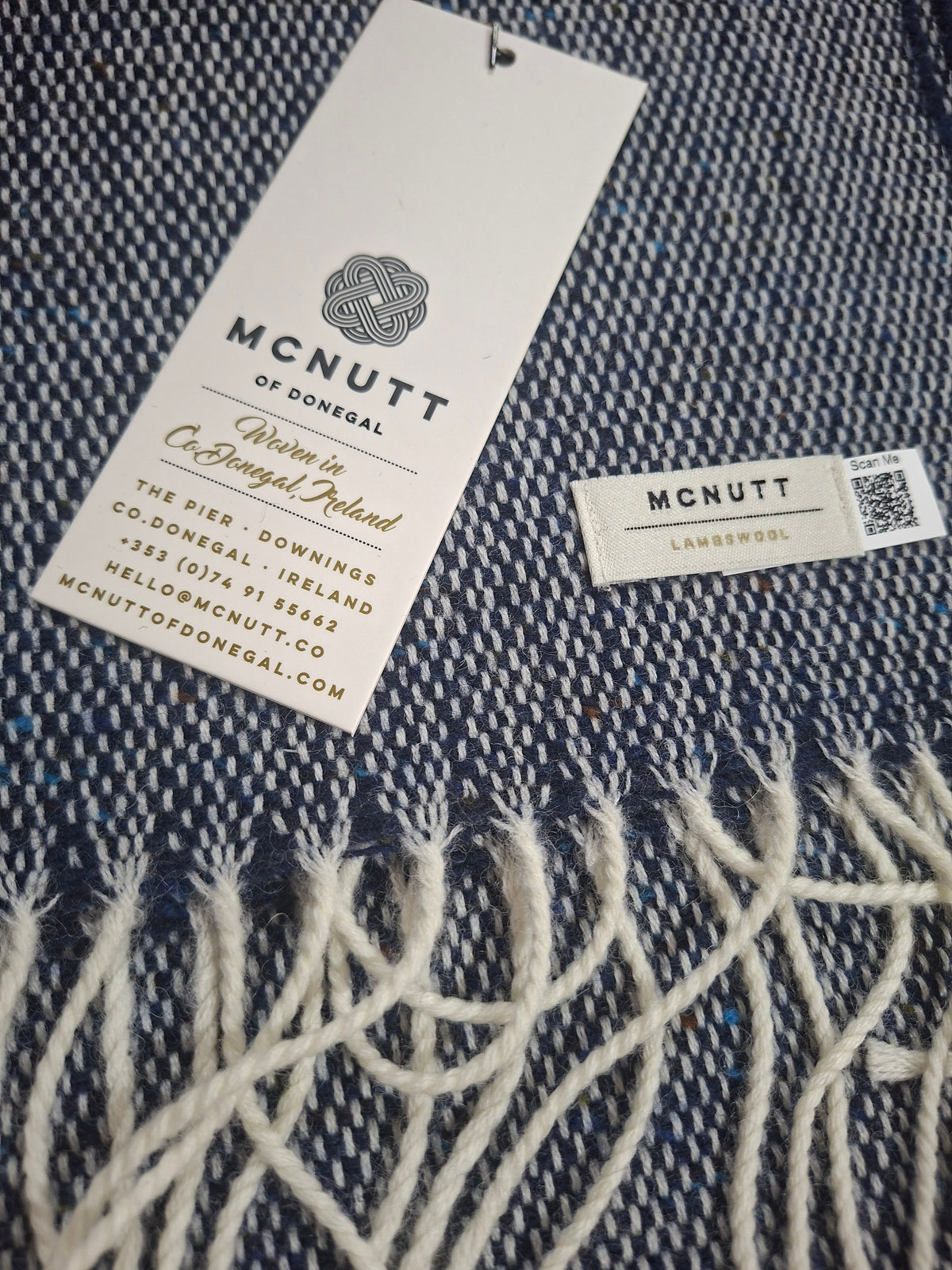 A close-up of lambswool navy scarf with cream fringe, showing a white brand tag that reads McNutt of Donegal and a small woven label.